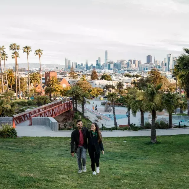 Una coppia cammina verso la telecamera con Dolores Park e lo skyline di San Francisco dietro di loro.